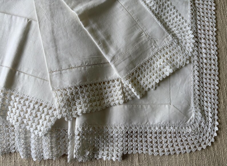 K&ouml;nnte beinhalten: Wei&szlig;e Bettw&auml;sche aus Leinen mit aufwendiger H&auml;kelspitze. Die Bettw&auml;sche ist gefaltet und zeigt das filigrane Lochmuster entlang der Kanten. Der Stoff wirkt weich und leicht strukturiert, was auf Vintage oder Handarbeit hindeutet.