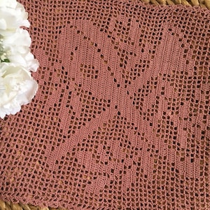 May include: A dusty rose crocheted doily with a delicate, intricate pattern. The doily is made with a fine yarn and has a square shape.