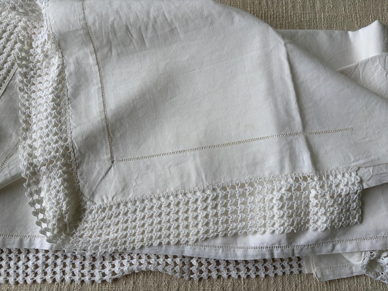 K&ouml;nnte beinhalten: Wei&szlig;es Leinen-Tischtuch mit geh&auml;keltem Spitzensaum. Das Tischtuch ist gefaltet und zeigt die Textur des Stoffes und die filigranen Details der Spitze. Die N&auml;hte sind an den R&auml;ndern sichtbar.