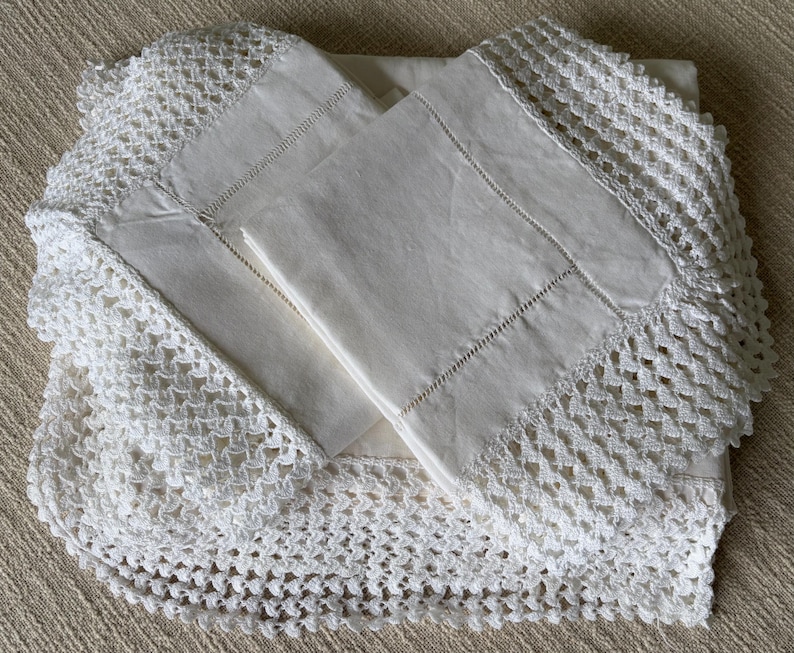 K&ouml;nnte beinhalten: Ein gefaltetes wei&szlig;es Leinen-Bettw&auml;sche-Set mit aufwendiger H&auml;kelspitze. Das Set enth&auml;lt Kissenbez&uuml;ge und ein Laken, alles in einer neutralen Farbe. Die Spitzendetails verleihen der Bettw&auml;sche eine Vintage- und handwerkliche &Auml;sthetik.