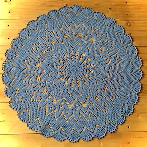 May include: A round, blue crocheted doily with a detailed floral and geometric pattern. The doily is made of a soft, textured yarn and has a scalloped edge. The handmade piece is displayed on a wooden surface.