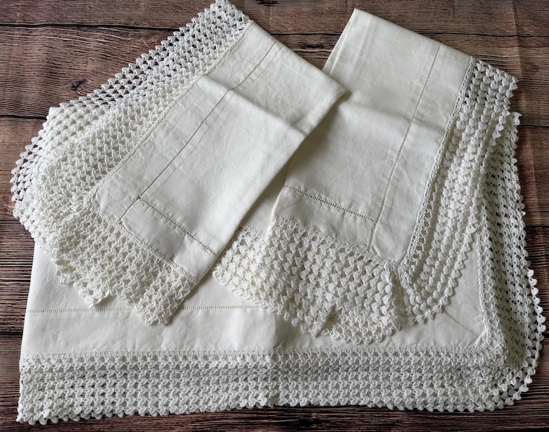 K&ouml;nnte beinhalten: Cremefarbene Bettw&auml;sche mit aufwendiger H&auml;kelspitze. Das Set beinhaltet ein Bettlaken und zwei Kissenbez&uuml;ge, alle ordentlich gefaltet. Die H&auml;kelarbeit verleiht der Bettw&auml;sche eine zarte, vintage &Auml;sthetik.