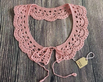 May include: A delicate pink crocheted collar with a tie closure. The collar is made with a lacy pattern and has a handmade tag that says "Handmade with love".