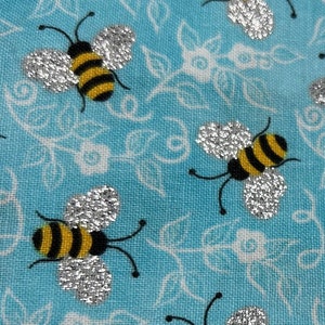 Puede incluir: Una tela azul con un patrón repetido de abejas de purpurina plateada sobre un diseño floral blanco.