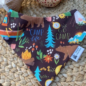 May include: A brown triangular bandana with a camping theme. The bandana features a variety of camping-related images, including a campfire, a tent, a compass, a canoe, a bear, and a pine tree. The bandana also has the text "Camp Life" and "Go Camp" printed on it. The bandana is made of cotton fabric and has a white paw print with the text "Claws N Paws Shop" printed on it.