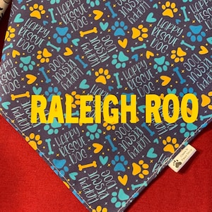 Puede incluir: Un pañuelo morado con un patrón de huellas de patas y huesos. El pañuelo tiene el texto "Happy Rescue Dog" repetido en todo el pañuelo. El texto "RALEIGH ROO" está impreso en amarillo en el pañuelo.