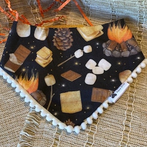 May include: A black dog bandana with a white pom-pom trim, featuring a repeating pattern of s'mores, campfires, and pinecones. The bandana has the text "Paws Shop" on a white label.