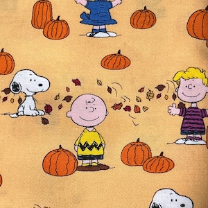 May include: A light orange fabric featuring a repeating pattern of Peanuts characters including Charlie Brown, Lucy, and Snoopy. The design includes orange pumpkins and scattered brown, red, and yellow autumn leaves across the entire surface.