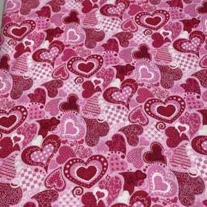 May include: Pink fabric with a repeating pattern of hearts in various shades of pink and red. The hearts have different designs, including solid, dotted, and patterned textures. The fabric is suitable for crafts and sewing projects.