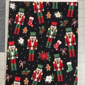 May include: Black fabric with a repeating Christmas pattern. The design includes nutcrackers in red and green uniforms, gingerbread men, poinsettias, candy canes, and Christmas stockings. White dots are scattered across the background.