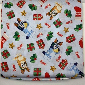 May include: Light blue fabric featuring a repeating pattern of cartoon dogs, Christmas presents, holly, candy canes, stars, and red stockings. The fabric is folded, showing the pattern on the front.