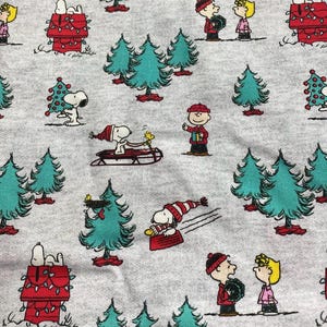 May include: A gray fabric featuring a repeating pattern of Snoopy, Charlie Brown, and other Peanuts characters in a winter scene. The design includes red doghouses, Christmas trees, and characters sledding.