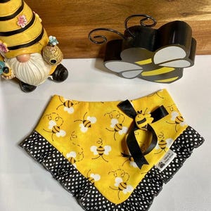 May include: A yellow dog bandana with a bee pattern and black polka dot trim. The bandana has a black bow and a bee-shaped embellishment. A gnome figurine and a bee-shaped decoration are in the background.