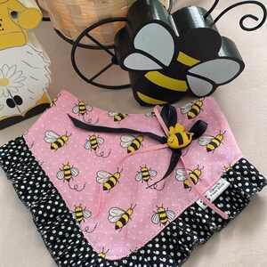 May include: Pink pet bandana with a bee pattern and black polka dot trim. The bandana has a black ribbon and a yellow bee embellishment. A decorative bee and a basket are in the background.