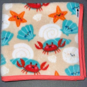 May include: A beige fleece blanket with a coral border features a repeating pattern of blue seashells, red crabs, and orange starfish. The blanket has a small white tag with the text "Cozy & Paws" on it.