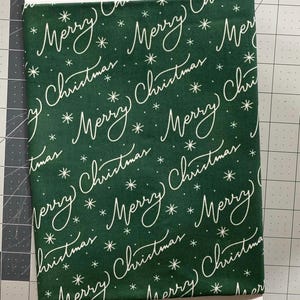 May include: A folded piece of fabric with a dark green background. The fabric is patterned with the words "Merry Christmas" in a white, cursive font, along with small white snowflake designs. The fabric is likely for holiday crafts.