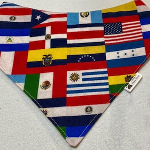 May include: A triangular bandana with a patchwork design of various flags from around the world. The bandana is made of a soft, cotton fabric and features a variety of colors, including red, white, blue, yellow, and green. The bandana is trimmed with a white stitching and has a small tag attached to the bottom right corner that reads "Class II Paws Shop".