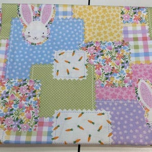Peut inclure: Un tissu patchwork sur le thème du printemps. Le motif présente des carrés de différents motifs, notamment des imprimés floraux, des pois et du vichy. Des visages de lapins et des motifs de carottes sont incorporés dans le design. La palette de couleurs comprend des tons pastel de bleu, rose, jaune et vert.
