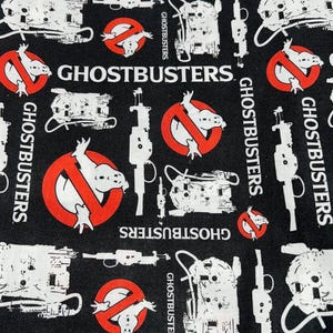 May include: Black fabric with a repeating pattern of the Ghostbusters logo, a ghost in a circle with a red slash through it, and a proton pack. The text "Ghostbusters" appears in white.
