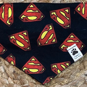 May include: Black bandana with a repeated pattern of red and yellow Superman logos. The bandana has a white tag with the text "Claws N Paws Shop".