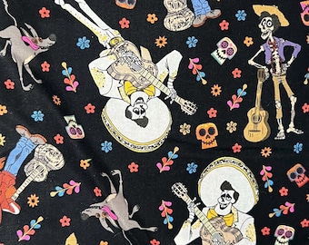 Op de afbeelding: Een zwarte stof met een herhalend patroon van personages, waaronder een jongen in een rode hoodie, skeletmuzikanten in witte mariachi-pakken met gitaren en een grijze hond, versierd met kleurrijke schedels en kleine bloemmotieven.