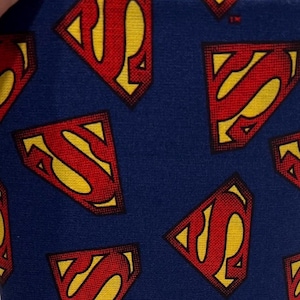 May include: A close-up of blue fabric patterned with the Superman logo. The logo is a red 'S' within a yellow and red shield. The fabric has a textured appearance, with the logos repeating across the surface.