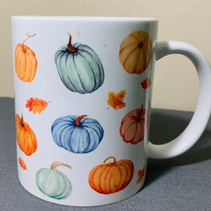 May include: White ceramic mug decorated with watercolor pumpkins in shades of orange, blue, and green, along with autumn leaves. The mug has a curved handle and is suitable for hot or cold beverages.