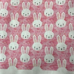 May include: Pink fabric with a repeating pattern of white bunny faces and ears. The bunnies are set against a pink background with horizontal stripes. The bottom edge has a white border.