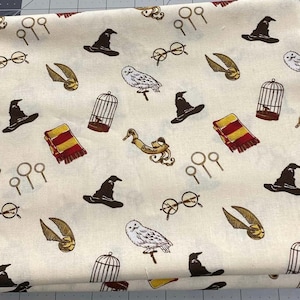 May include: Cream-colored fabric with a repeating pattern of wizarding-themed illustrations. The design includes black wizard hats, golden snitches, owls, round glasses, scarves, and birdcages. The fabric is folded, showing the pattern's repeating nature.