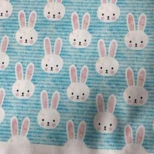 May include: Fabric with a repeating pattern of white bunny faces with pink ears on a light blue background. The bunnies have black eyes and noses. The fabric has a textured appearance.