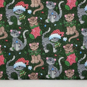 May include: A green fabric with a repeating pattern of gray and brown cats wearing Santa hats, scarves, and Christmas decorations. The cats are interspersed with snowflakes and Christmas lights. The fabric has a white border.