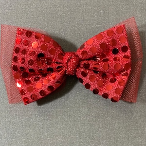 May include: A red sequined bow with a tulle overlay. The bow is a vibrant red color and features large, round sequins. The tulle adds a delicate texture to the bow. This accessory is suitable for hair or clothing.