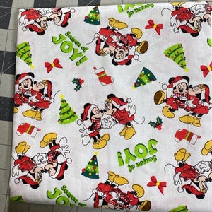Peut inclure: Tissu blanc orné de Mickey et Minnie Mouse en tenues de Noël, avec des sapins verts, des nœuds rouges et des bas de Noël. Les mots "Season of JOY!" sont imprimés en vert.