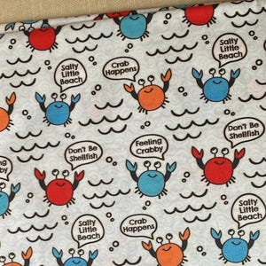 May include: White fabric with a repeating pattern of cartoon crabs in red, orange, and blue. The crabs are interspersed with wavy lines and speech bubbles with phrases like "Salty Little Beach" and "Crab Happens".