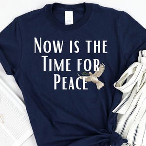 Könnte beinhalten: Marineblaues T-Shirt mit dem Text "Now is the Time for Peace" und einer Taubengrafik. Das Shirt wird mit einer gestreiften Hose und einer Zeitschrift mit Goldschmuck präsentiert.