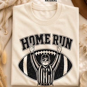 Puede incluir: Una camiseta de color natural con un estampado gráfico negro de un árbitro con los brazos levantados en señal de touchdown, frente a una forma de balón de fútbol. El texto "HOME RUN" aparece en una fuente negra, negrita y arqueada.