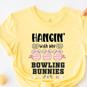 以下が含まれることがあります： 「Hangin' with my Bowling Bunnies」という文字と、イースターエッグを持ったウサギ3匹のイラストが描かれた黄色のTシャツです。