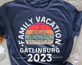 以下が含まれることがあります： 「ファミリーバケーション Gatlinburg 2023」と書いてあるヴィンテージスタイルのグラフィックがプリントされたネイビーブルーのTシャツ。グラフィックには、山と木々のある夕焼けが描かれています。