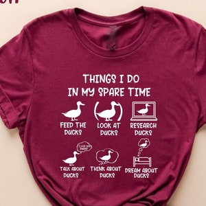 May include: A maroon t-shirt with white text that reads "Things I do in my spare time". The text is surrounded by illustrations of ducks, including feeding ducks, looking at ducks, researching ducks, talking about ducks, thinking about ducks, and dreaming about ducks.