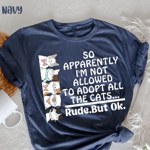 Può includere: Una maglietta blu navy con una grafica di una fila di gatti e il testo "SO APPARENTLY I'M NOT ALLOWED TO ADOPT ALL THE CATS... Rude.But Ok."