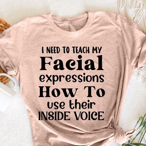 May include: A light pink t-shirt with black text that reads "I need to teach my Facial expressions How To use their INSIDE VOICE".