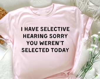 Może przedstawiać: Różowy T-shirt z nadrukiem "Mam selektywny słuch, przepraszam, nie zostałeś wybrany dzisiaj" w kolorze czarnym.