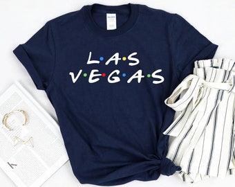 May include: A navy blue t-shirt with the text "L.A.S VEGAS" in white with colourful dots between the letters. The shirt is folded and laid on a white surface with a white and black striped skirt and a gold necklace.