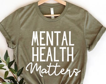 May include: A green t-shirt with the text "MENTAL HEALTH MATTERS" in white lettering. The shirt is on a hanger and has a knotted bottom.