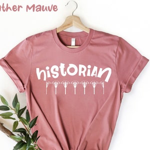 May include: Heather mauve t-shirt with the word "historian" in white text and a row of stylised white flowers below the word.