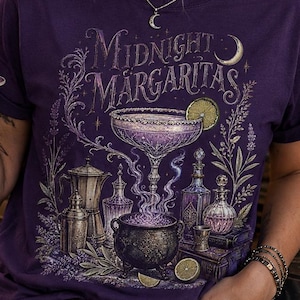 May include: A dark purple short-sleeve t-shirt featuring a graphic print with the text Midnight Margaritas. The design includes a steaming cauldron, a cocktail glass, various glass bottles, and lime slices, all rendered in shades of purple and white.