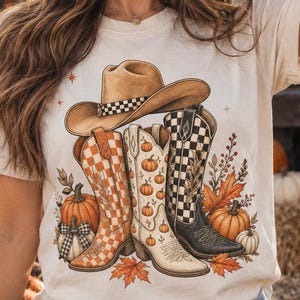 Può includere: Una t-shirt color crema con una stampa grafica di un cappello da cowboy beige appoggiato sopra tre stivali in stile western. Gli stivali presentano motivi a scacchi arancioni, disegni di zucche e scacchi bianchi e neri, circondati da foglie autunnali e zucche.