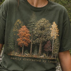 May include: An olive green short-sleeved t-shirt featuring a detailed graphic print of a forest with various tree species in shades of orange, yellow, and green. The text "easily distracted by trees" is printed in a simple font below the graphic.