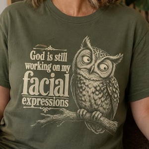 Puede incluir: Una camiseta de manga corta color verde oliva con el gráfico de un búho posado en una rama. El texto dice "God is still working on my facial expressions". El diseño está impreso en un color crema claro en la parte frontal de la prenda.