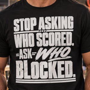 May include: A black short-sleeved crew neck t-shirt featuring white distressed-style text that reads STOP ASKING WHO SCORED. ASK WHO BLOCKED. The graphic is centered on the chest with horizontal lines above and below the text.